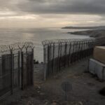 Ein massiver Grenzzaun mit Stacheldraht am Hafen von Ceuta bei düsterem Himmel, mit verrostetem Wachturm und Stapel von