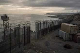 Ein massiver Grenzzaun mit Stacheldraht am Hafen von Ceuta bei düsterem Himmel, mit verrostetem Wachturm und Stapel von